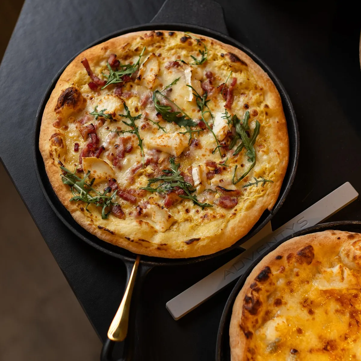 Zwei Pizzen in Pizzapfannen auf einem Tisch mit einer Flasche scharfer Soße im Hintergrund