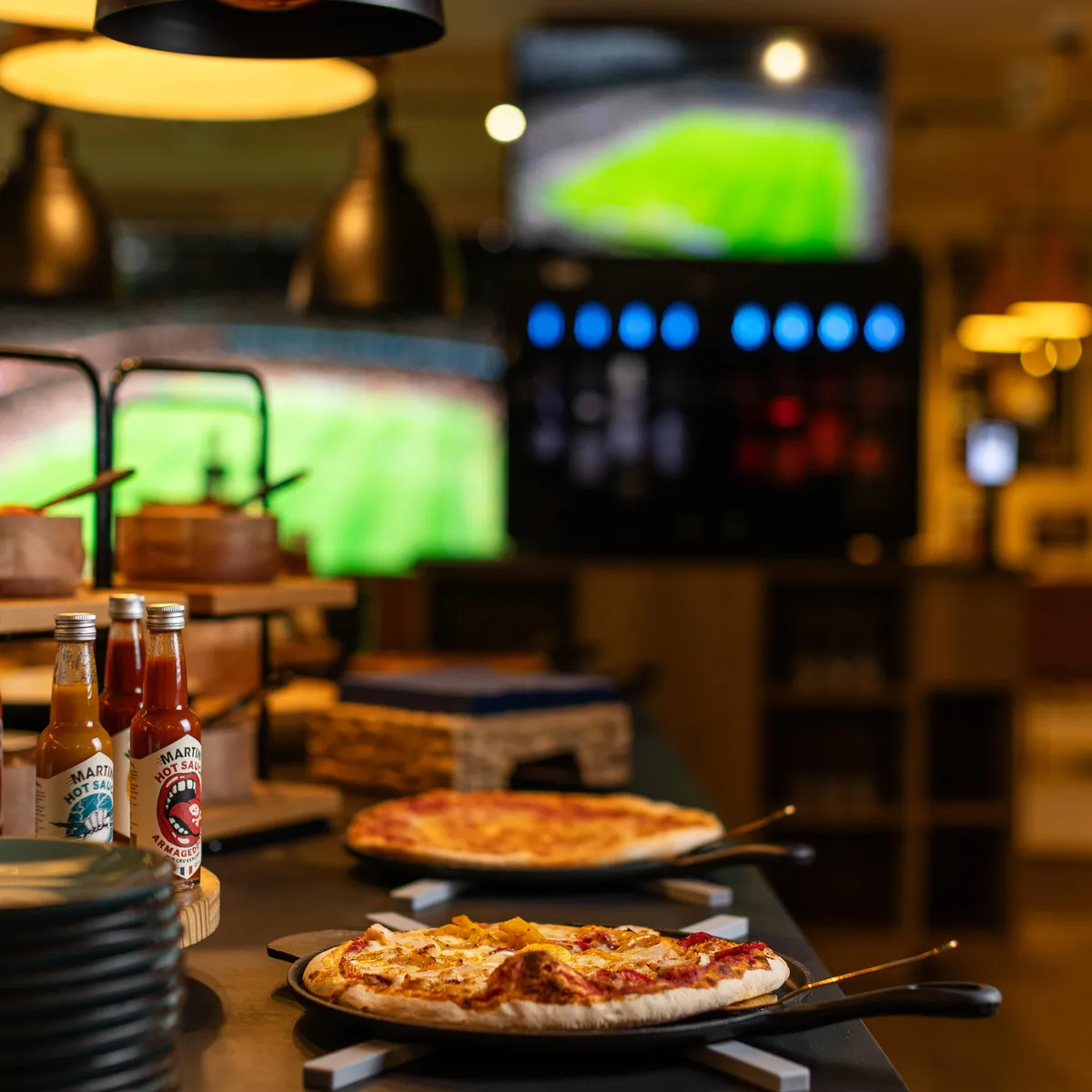 Zwei Pizzen auf Metallblechen, die auf einem Tisch in einem Restaurant stehen, mit im Hintergrund laufenden Bildschirmen, die ein Fußballspiel übertragen.
