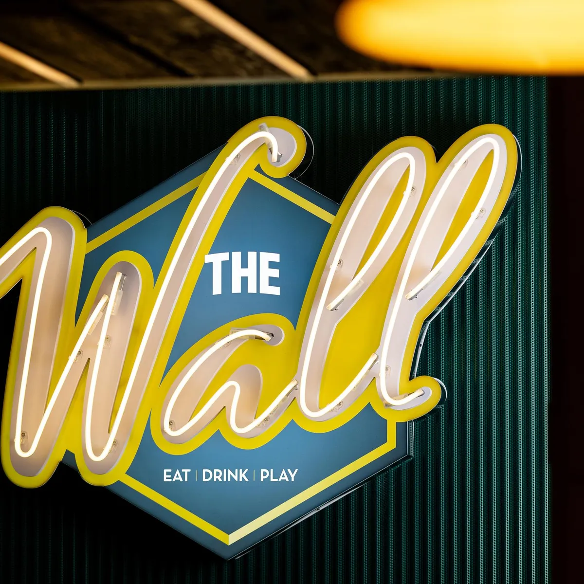 Leuchtende Schilder mit der Aufschrift "The Wall" und den Worten "Eat Drink Play" darunter, an einer dunklen Wand befestigt