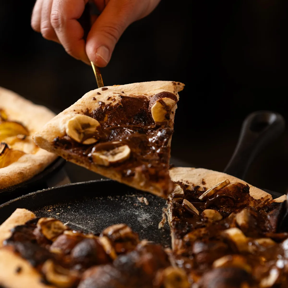 Eine Hand hält eine Scheibe Pizza mit Pilzen und Nüssen, während zwei Pizzen auf Backblechen liegen.