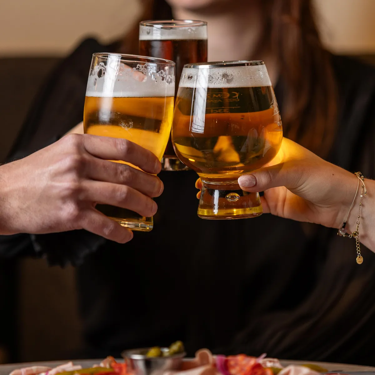 Zwei Personen erheben sich gegenüberstehend Biergläser, mit einem Teller mit Aufschnitt und Käse auf dem Tisch vor ihnen.