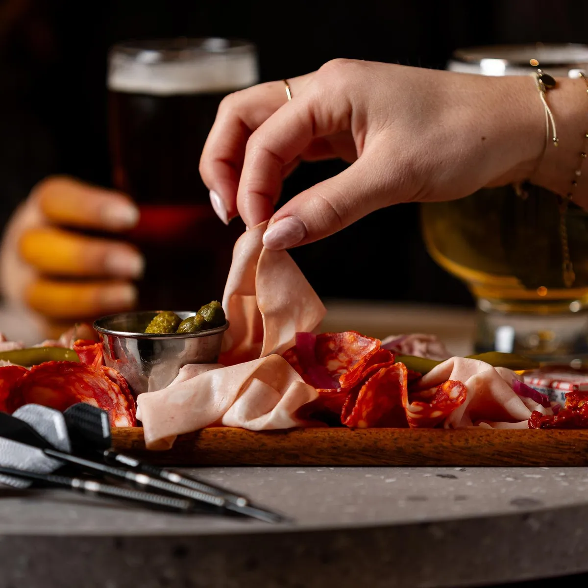 Eine Person nimmt ein Stück Fleischwurst von einem Brett mit Gurken und Oliven, begleitet von Biergläsern.