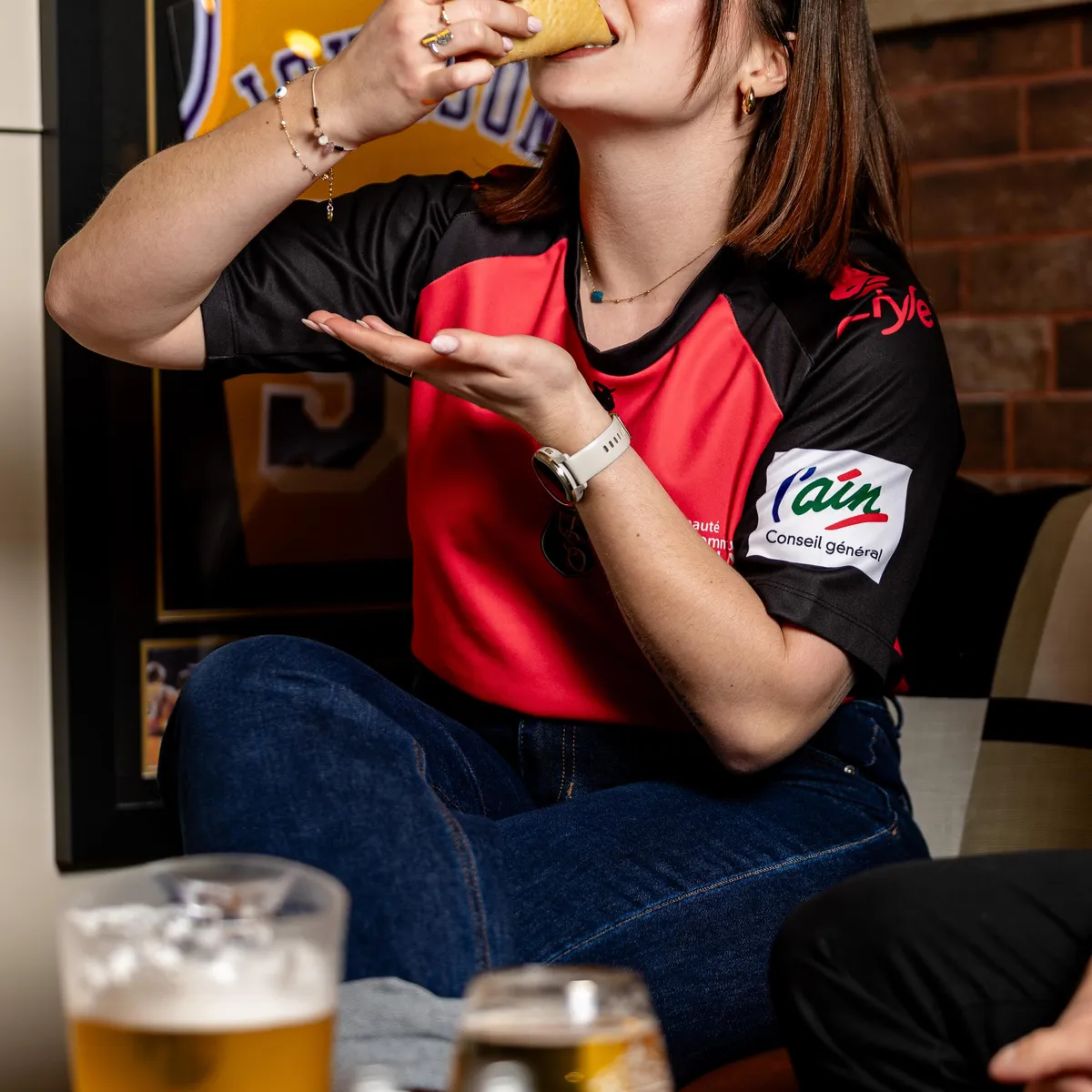 Eine Person isst ein Sandwich und hält ein Bierglas vor einem Fernseher.