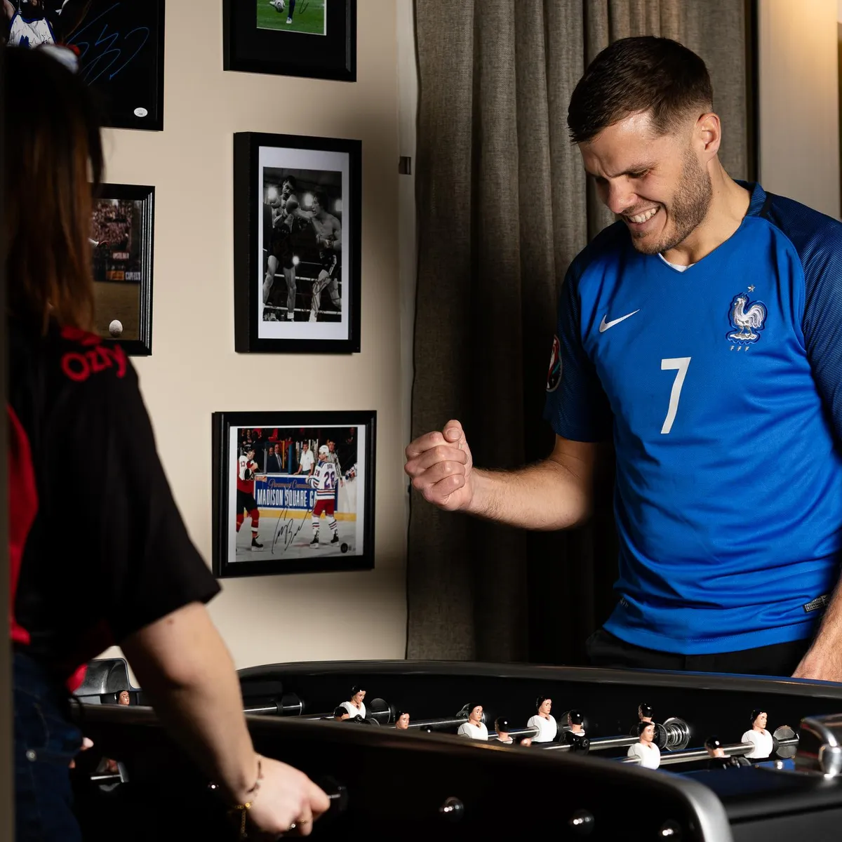 Ein Mann im blauen Fußballtrikot spielt mit zwei Frauen an einem Kickertisch in einem Raum mit gerahmten Fotos.
