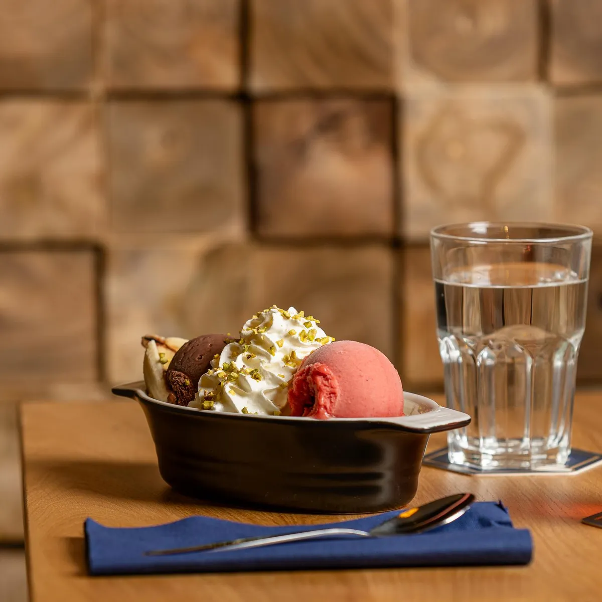 Eine Schüssel mit Beilagen, Eis und Toppings, ein Glas Wasser und ein Glas rotes Getränk auf einem Holztisch vor einer Backsteinwand