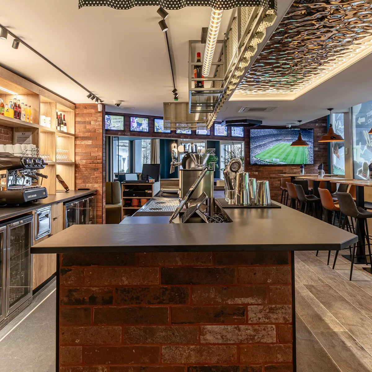 Bar mit zentraler Theke, Bildschirme mit einem Fußballspiel, Regale mit Flaschen und Barhockern
