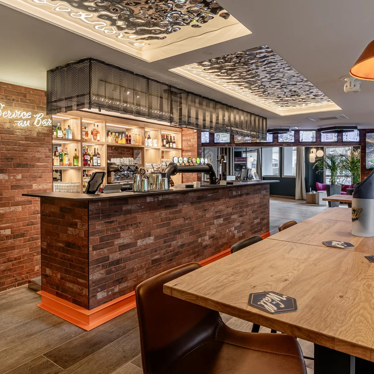 Bar mit Theke aus Ziegelsteinen, Regalen für Flaschen und Holztischen, mit Bildschirmen, die im Hintergrund ein Fußballspiel übertragen