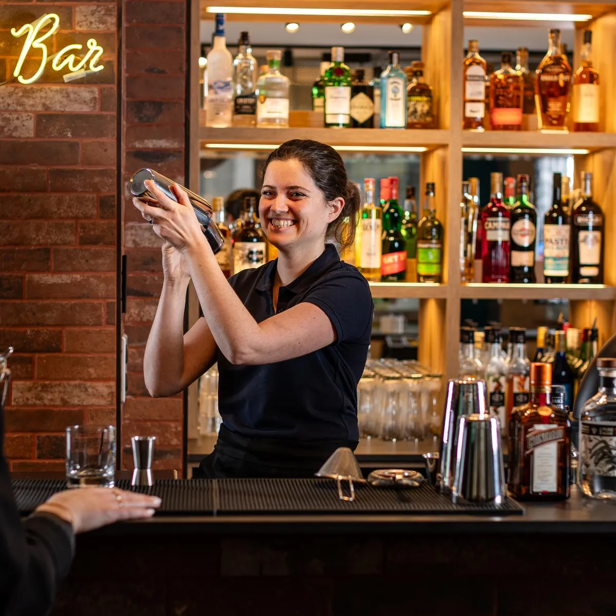 Eine Barkeeperin bereitet hinter einer gut bestückten Bar mit Flaschen und Spirituosen einen Cocktail zu.