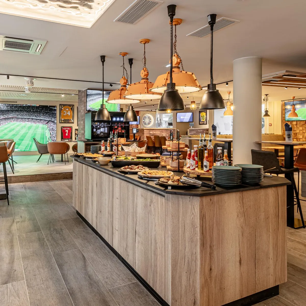 Eine Bar mit einer Theke aus hellem Holz, Hockern und Bildschirmen, die ein Fußballspiel übertragen, mit Getränken und Essen auf der Theke