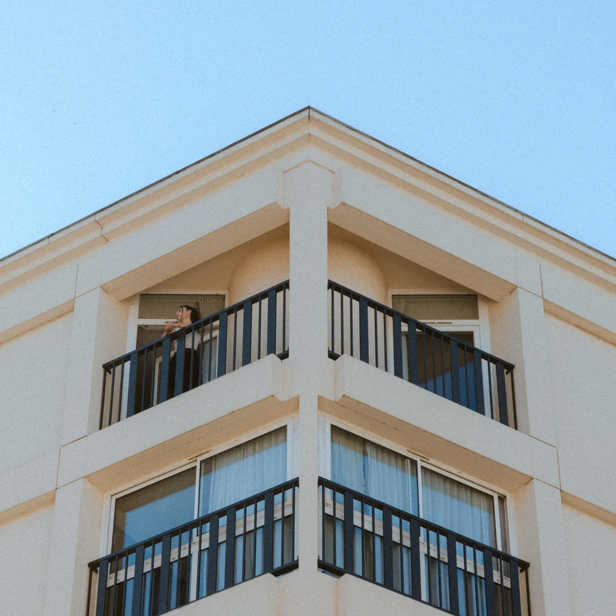 Person steht auf einem Balkon an der Ecke eines beigen Gebäudes mit schwarzen Balustraden