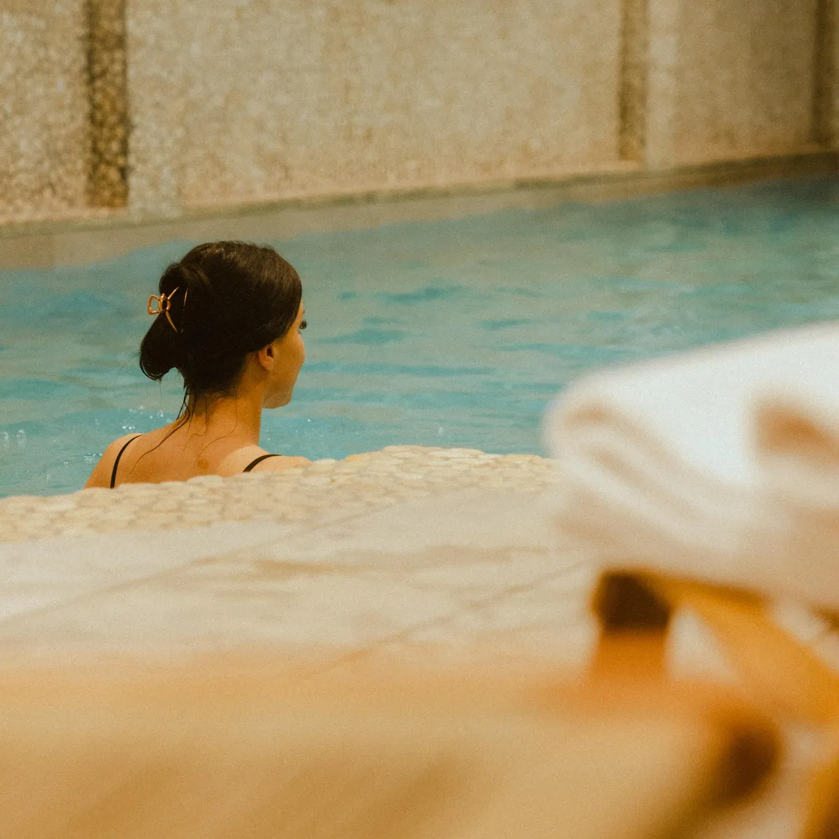 Une personne avec les cheveux attachés est assise dans une piscine, vue de dos, avec un mur en pierre en arrière-plan