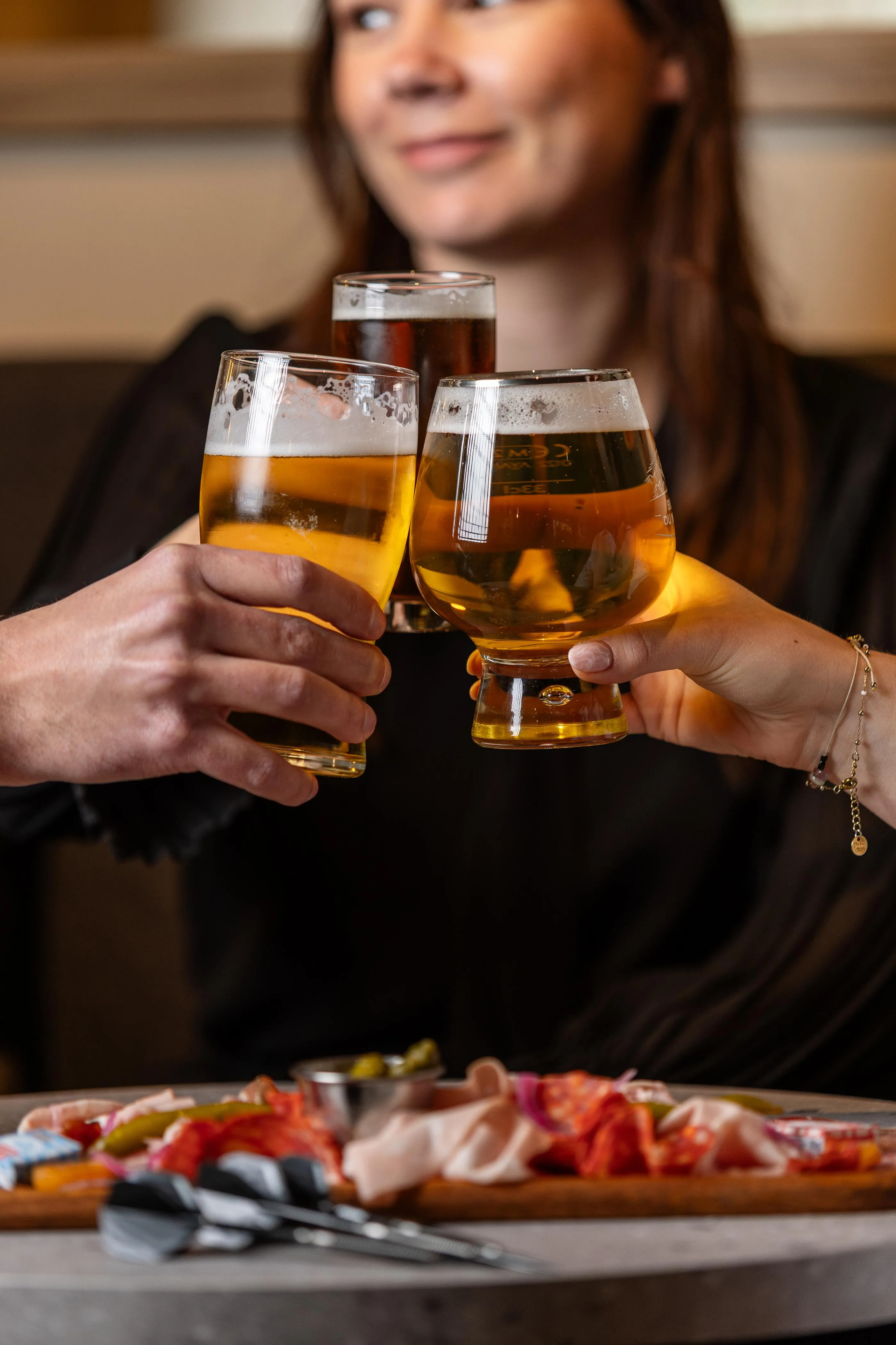 Deux personnes lèvent des verres de bière en se faisant face, avec une assiette de charcuterie et de fromage sur la table devant eux