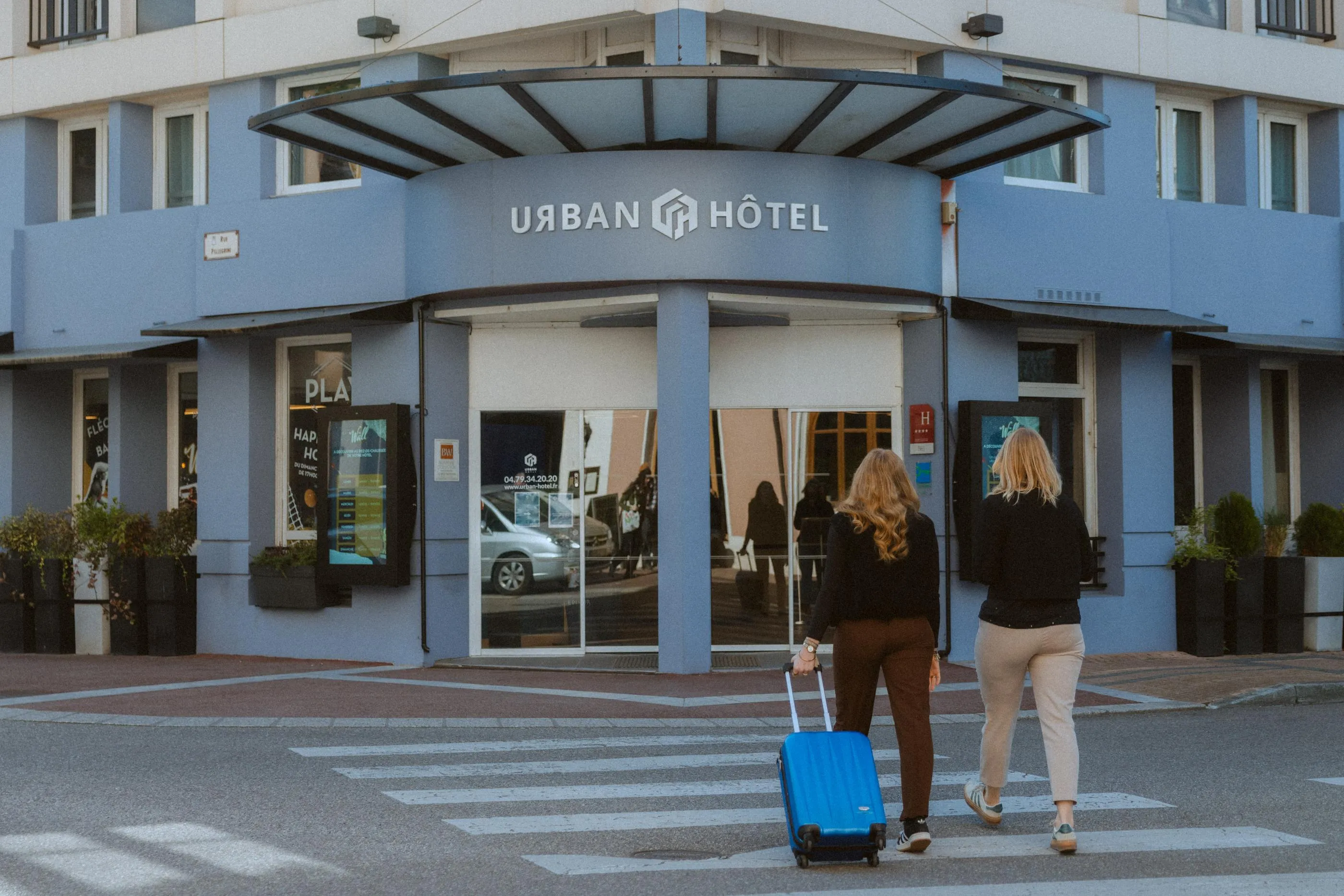 Zwei Personen schieben einen blauen Gepäckwagen und überqueren einen Fußgängerüberweg vor dem Eingang eines Hotels namens Urban Hotel.