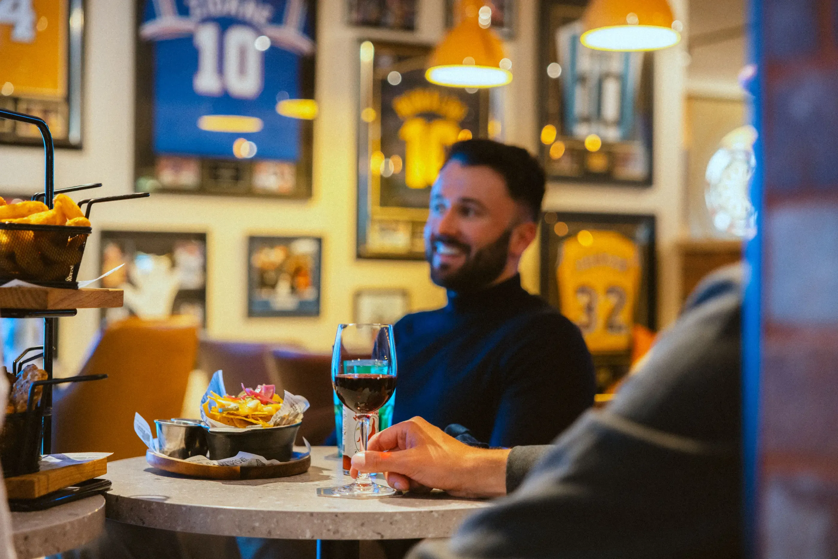 Ein Mann sitzt an einem Tisch mit einem Glas Wein und einer Schüssel Essen in einem mit Sportpostern dekorierten Restaurant.