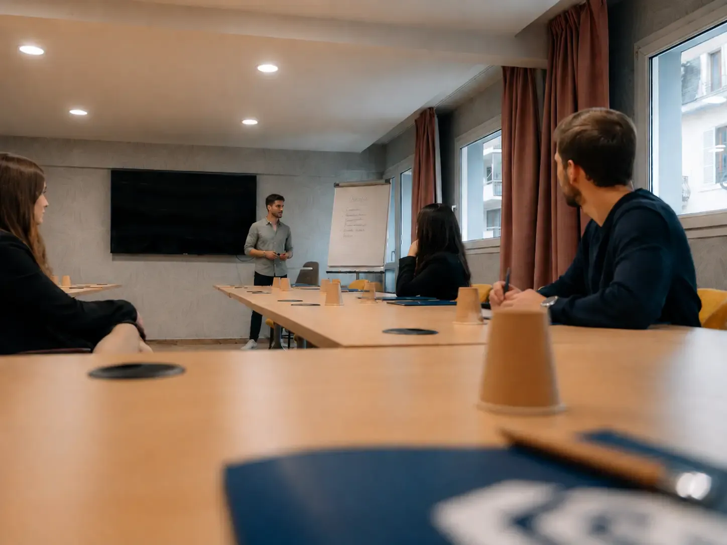 Vier Personen sitzen an einem Tisch, eine von ihnen steht vor einer Tafel und einem Bildschirm.