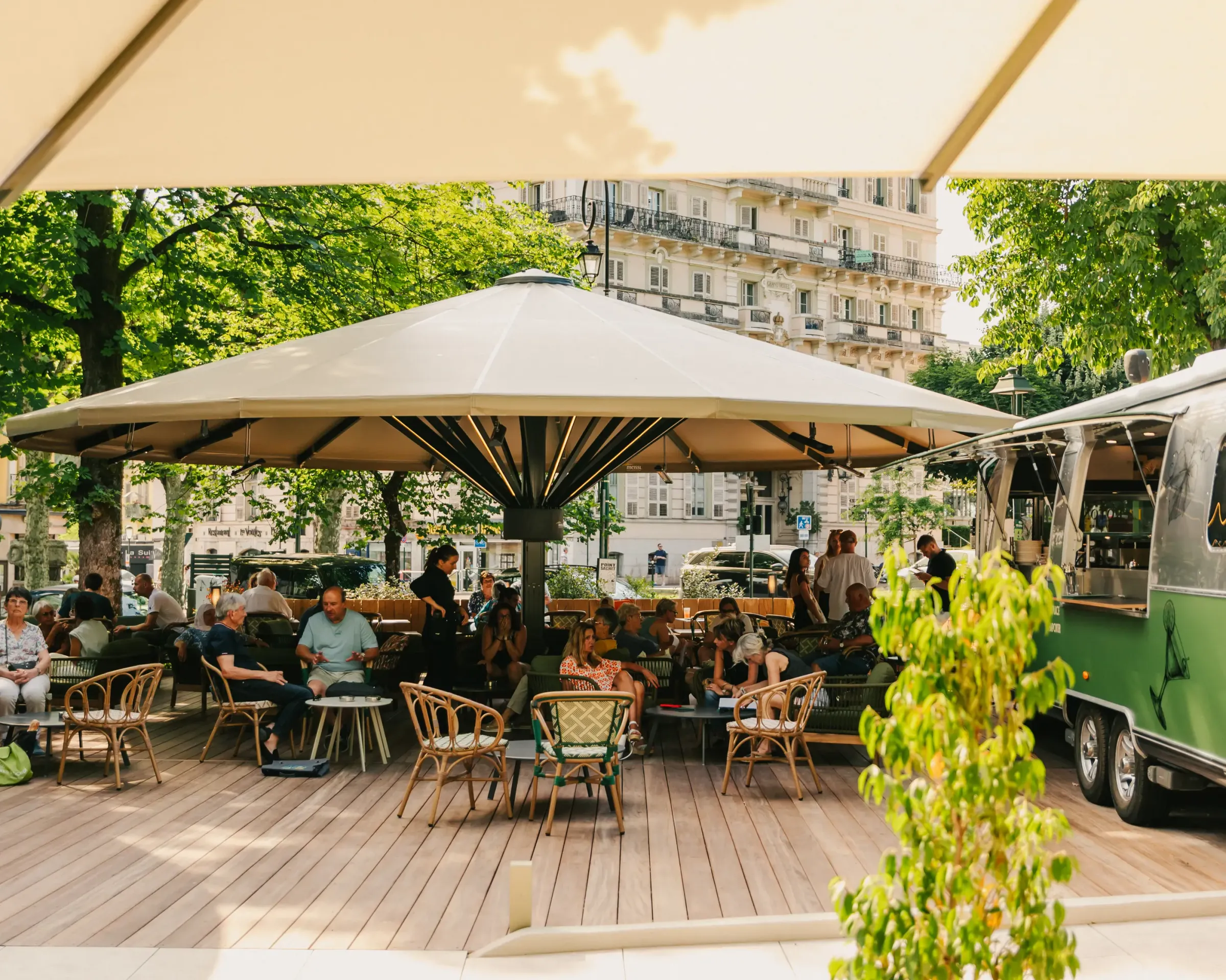 A covered terrace café with umbrellas, customers seated at tables under trees, surrounded by buildings and greenery.