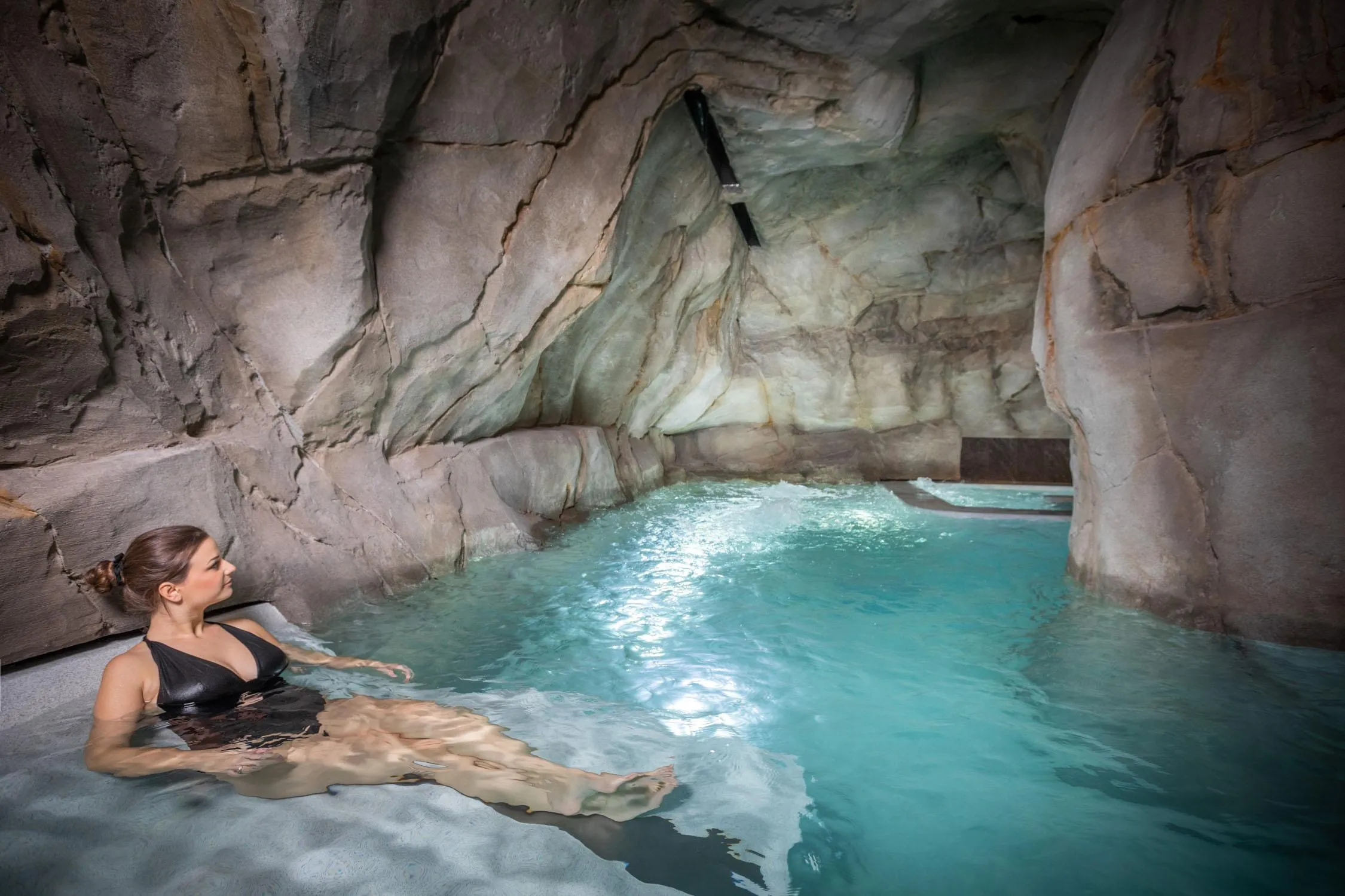 A person is lying in a blue water pool located in an artificial cave with rocky walls.