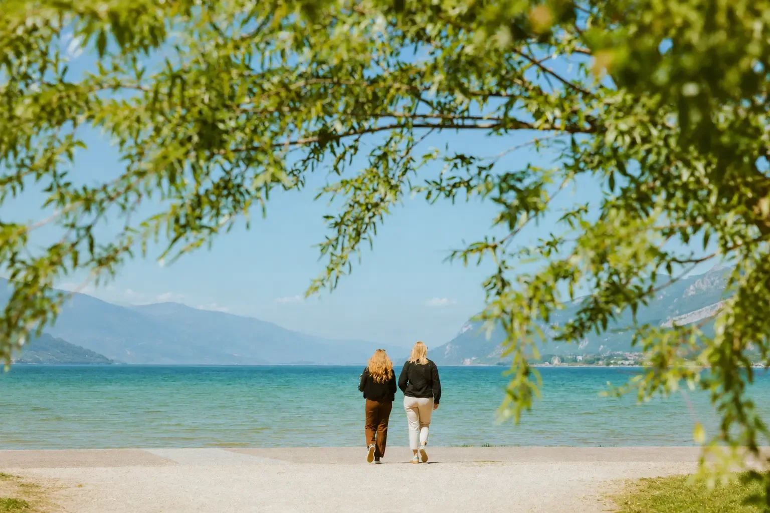 Two people are walking on a beach by a lake, with mountains in the background and tree branches in the foreground.